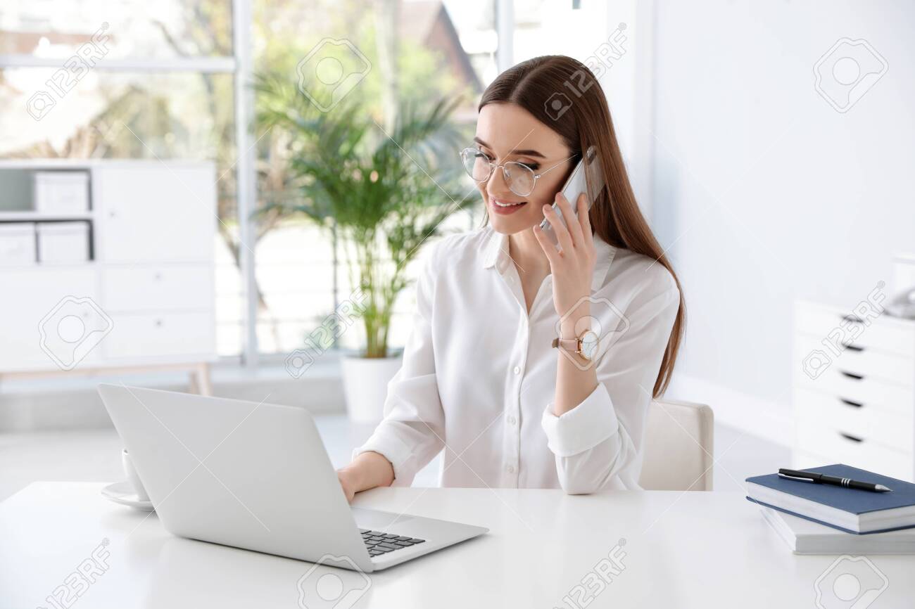 Young businesswoman talking on phone while using laptop at table in office - 133227094 Young businesswoman talking on phone while using laptop at table in office - 133227094