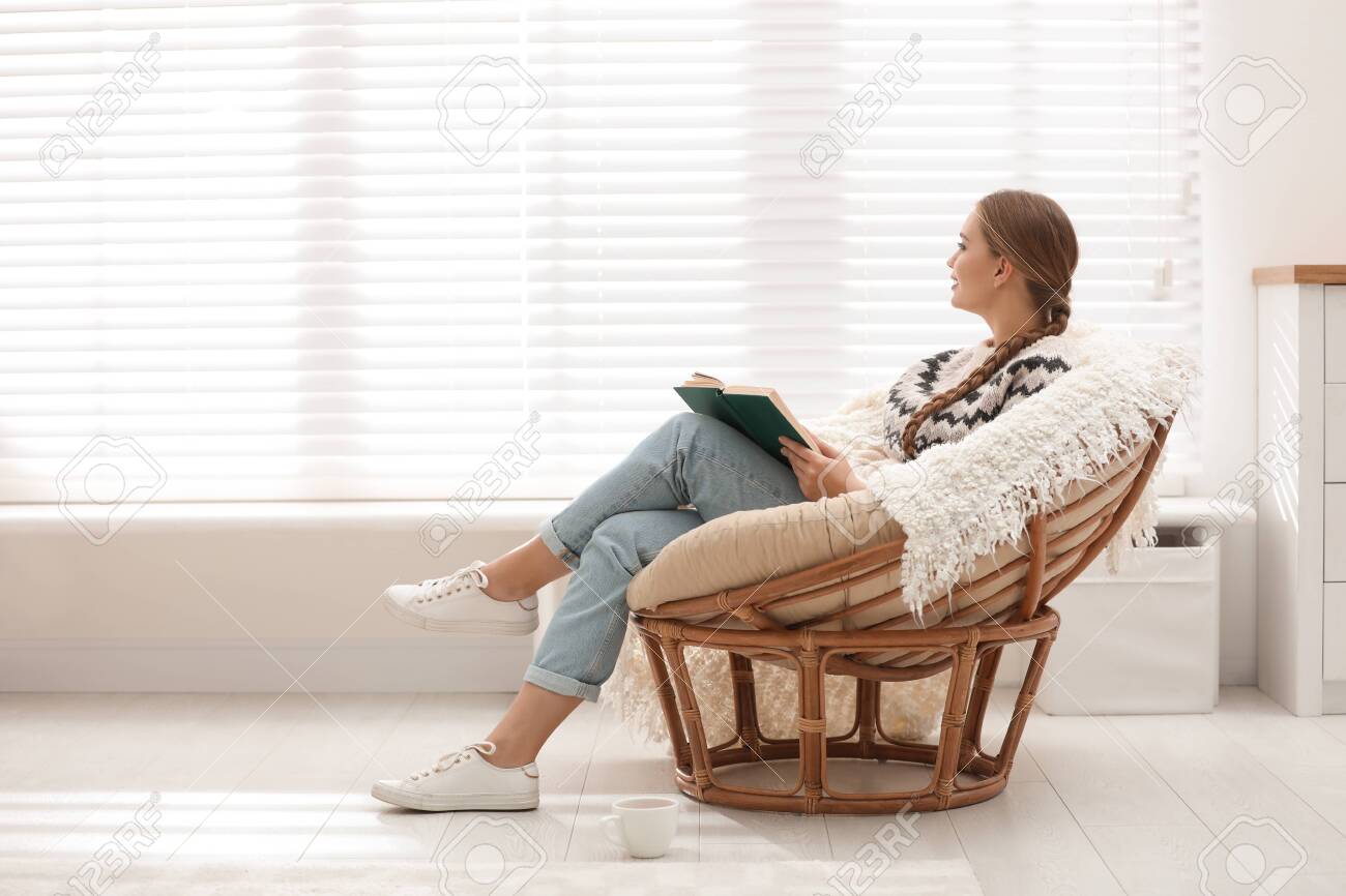 young woman reading book in papasan chair near window at home