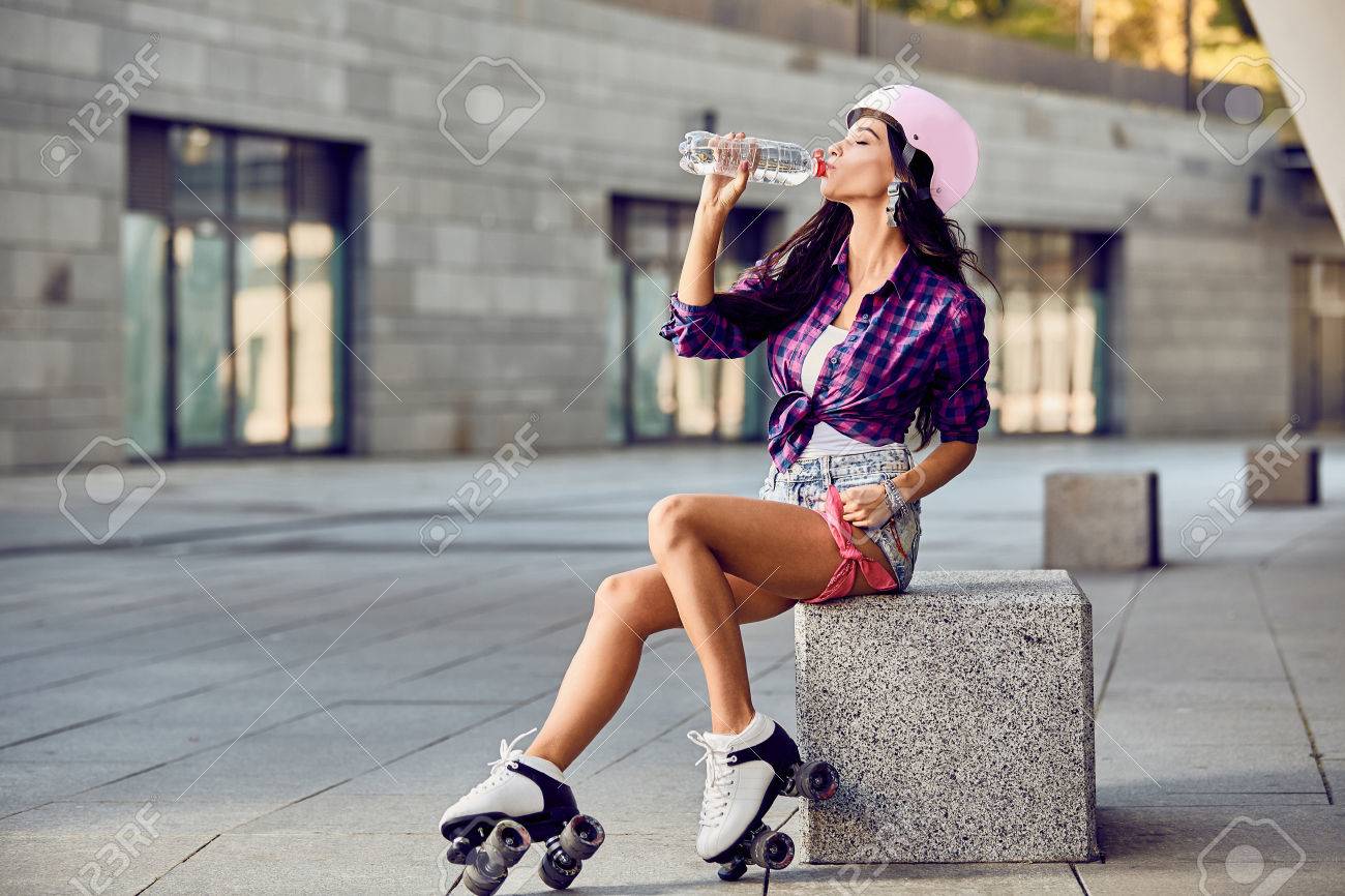 ローラー スケート ヘルメット飲料水の若い女性 ショート パンツ 紫のシャツ ローラーブレードでスタイリッシュな美しい少女 都市スケート公園でアクティブな時間後休んでいる内気な少女 の写真素材 画像素材 Image