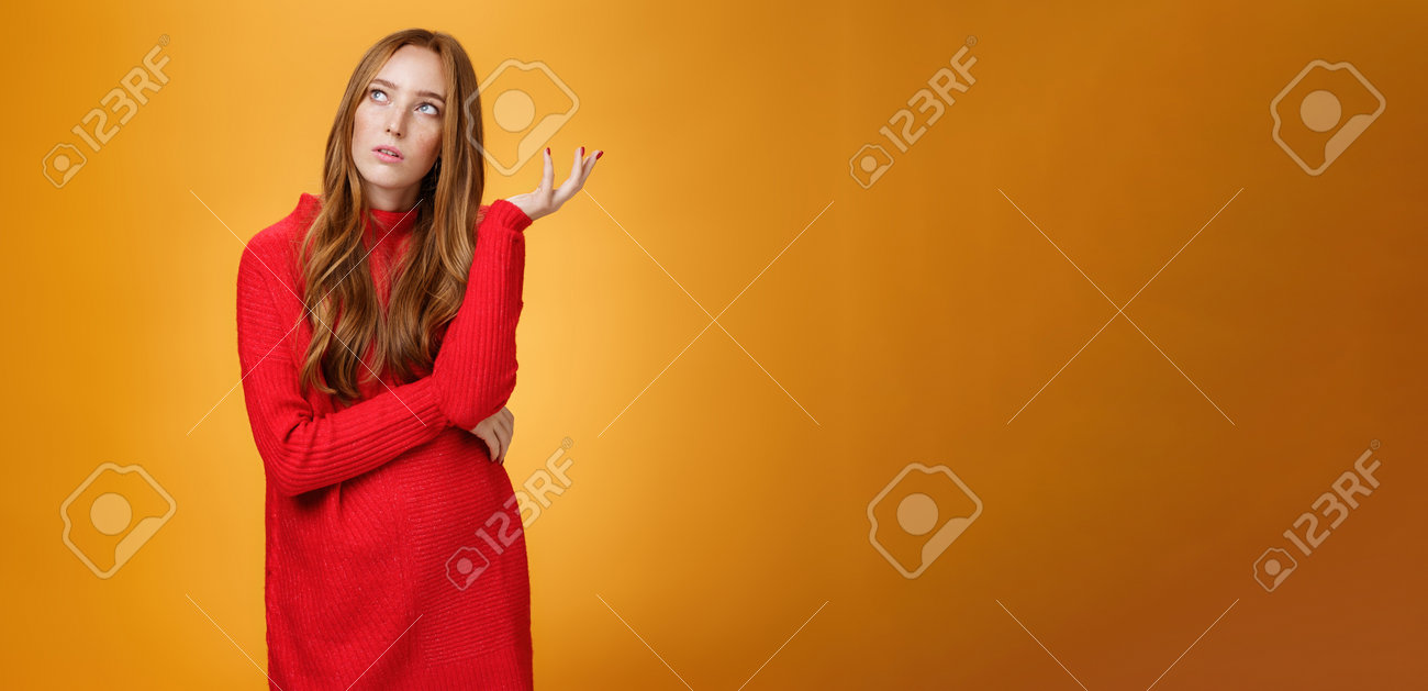 주황색 배경에 포즈를 취하면서 퍼즐을 푸는 향수를 불러일으키는 느낌을 떠올리며 왼쪽 상단 모서리에 불확실해 보이는 빨간 스웨터를 입은  불확실한 생각의 빨간 머리 여자친구의 초상화 로열티 무료 사진, 그림, 이미지 그리고 스톡포토그래피. Image