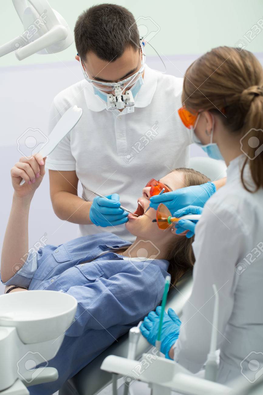 歯科用キャビネットで 患者用椅子で右手の鏡と青いシャツを着てかわいい女性患者 彼女の隣に男性歯科医とメスの助手があります 両方ブルー ラテックス手袋 青のマスクと白い制服を着用します また 歯医者を着用します の写真素材 画像素材 Image