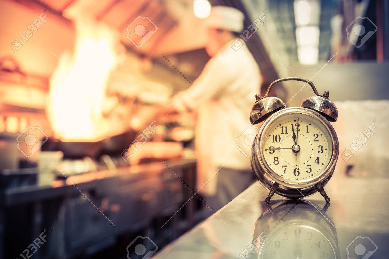 Alarm Clock With Blurred Chef Cooking In Kitchen Stock Photo