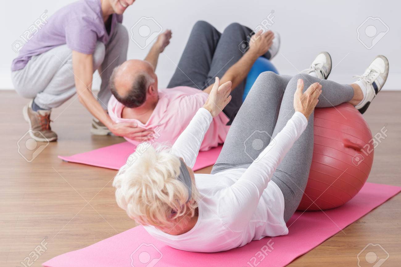 Senior exercising on gym ball with professional instructor. - 54417213 Senior exercising on gym ball with professional instructor. - 54417213