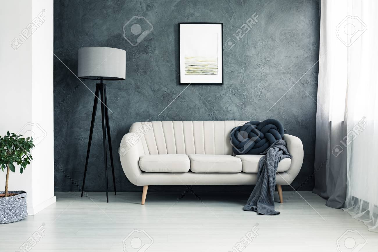 Bright couch with handmade knot cushion and grey blanket standing in a living room with lamp and poster hanging on textured wall - 90574868 Bright couch with handmade knot cushion and grey blanket standing in a living room with lamp and poster hanging on textured wall - 90574868
