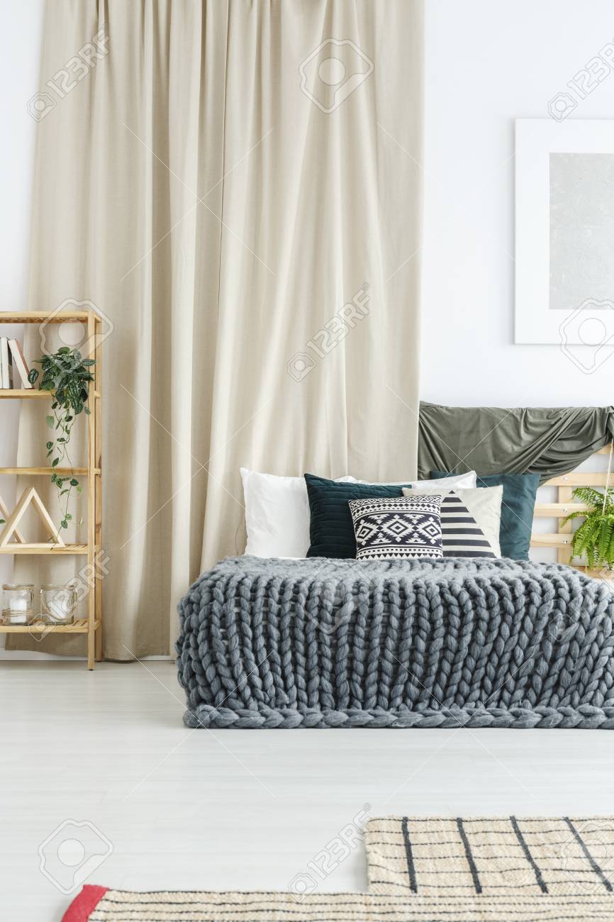 holzregal, kingsize-bett mit zierkissen und strickdecke vor beigem vorhang  im gemütlichen schlafzimmer