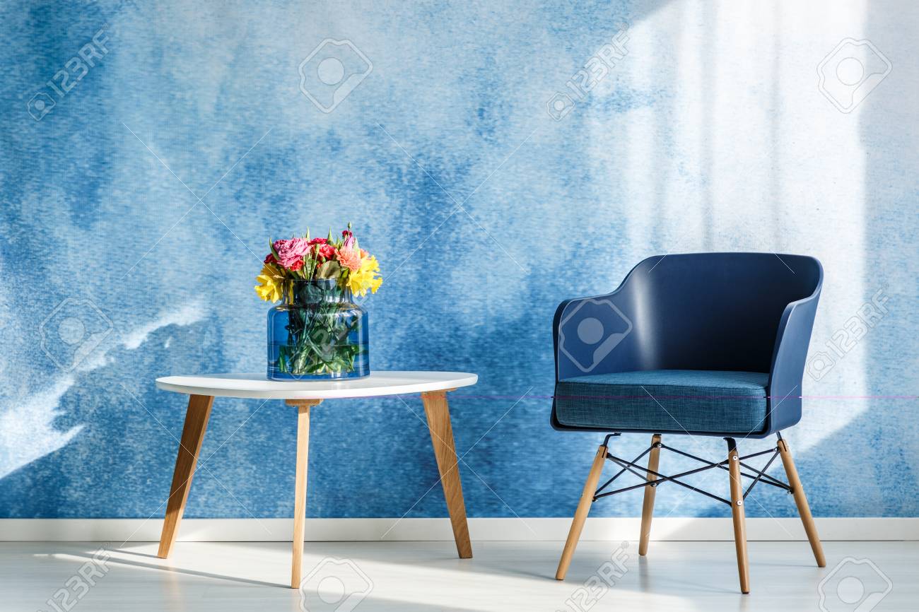 Colorful flowers on white wooden table next to navy blue armchair in simple living room interior