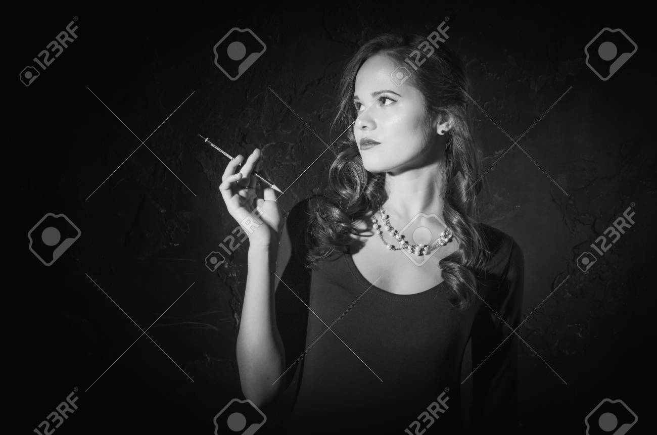ノワール映画スタイル女性長い黒のドレスとカメラ側に立って左手にタバコ 女性は手に鉛筆を使ってポーズをとって長い黒髪 黒と白の写真 古いファッション写真 の写真素材 画像素材 Image