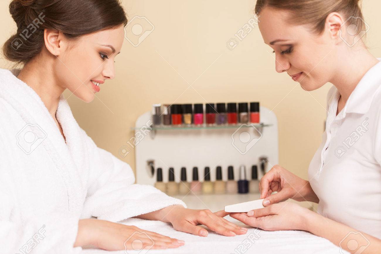 Close-up Of Beautician Hand Filing Nails Of Woman In Salon. two women smiling while sitting in spa salon - 33122091 Close-up Of Beautician Hand Filing Nails Of Woman In Salon. two women smiling while sitting in spa salon - 33122091
