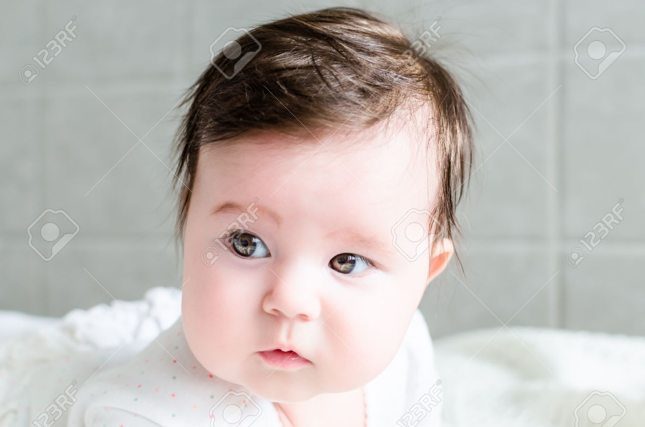 Retrato De La Nina Lindo Bebe Recien Nacido Dulce Morena Con El Pelo Negro En Blanco Agradable Manchado Traje De Mameluco Mirando Con Grandes Ojos Marrones Hermosos Castanos Y Mirada Curiosa En
