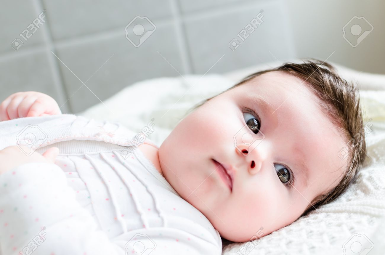 Retrato De La Nina Lindo Bebe Recien Nacido Dulce Morena Con El Pelo Negro En Blanco Agradable Manchado Traje De Mameluco Mirando Con Grandes Ojos Marrones Avellana Hermosa Miente En La Manta