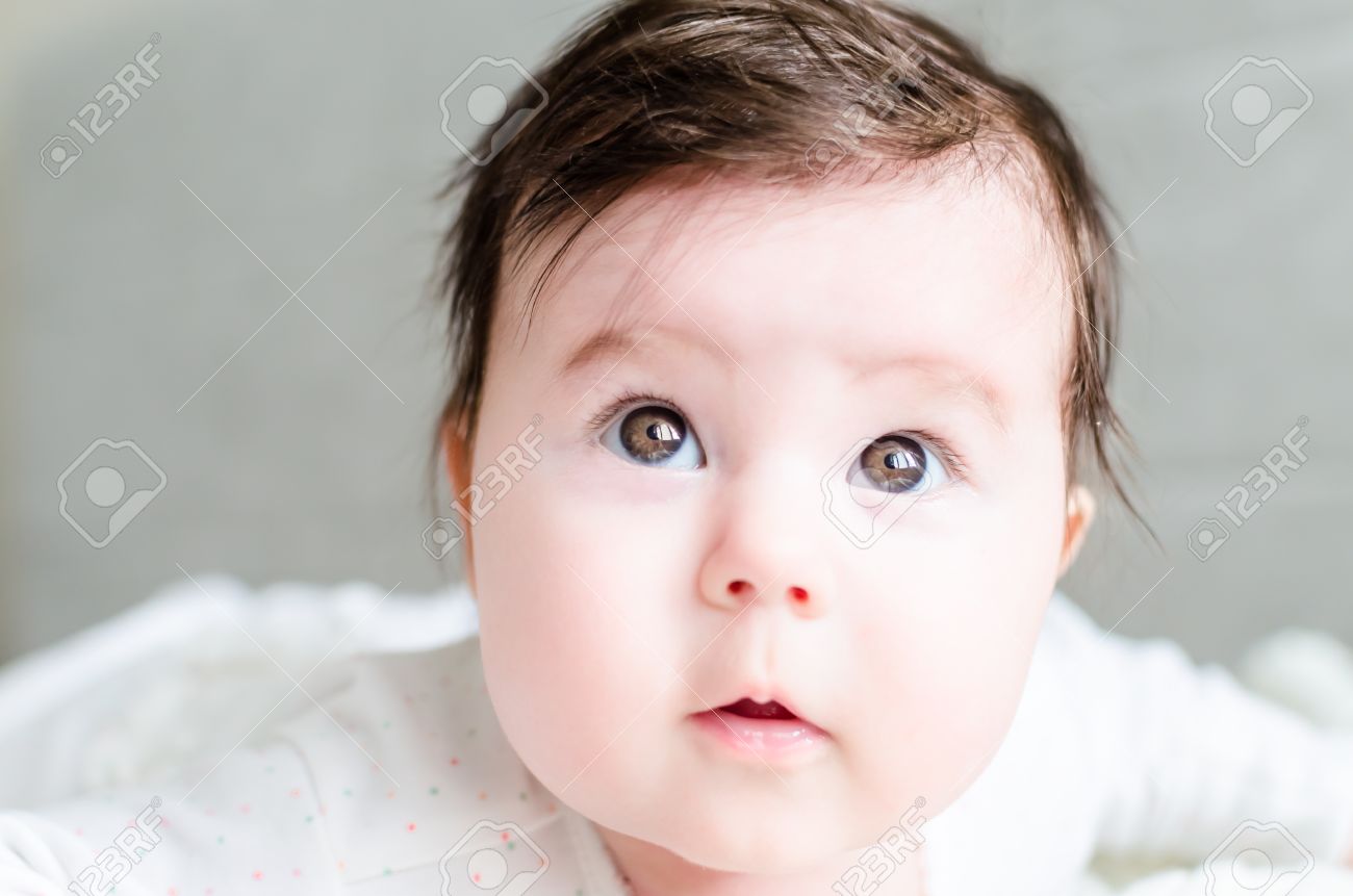 Retrato De La Nina Lindo Bebe Recien Nacido Dulce Morena Con El Pelo Negro En Blanco Agradable Manchado Traje De Mameluco Mirando Con Grandes Ojos Marrones Hermosos Castanos Y Mirada Curiosa En