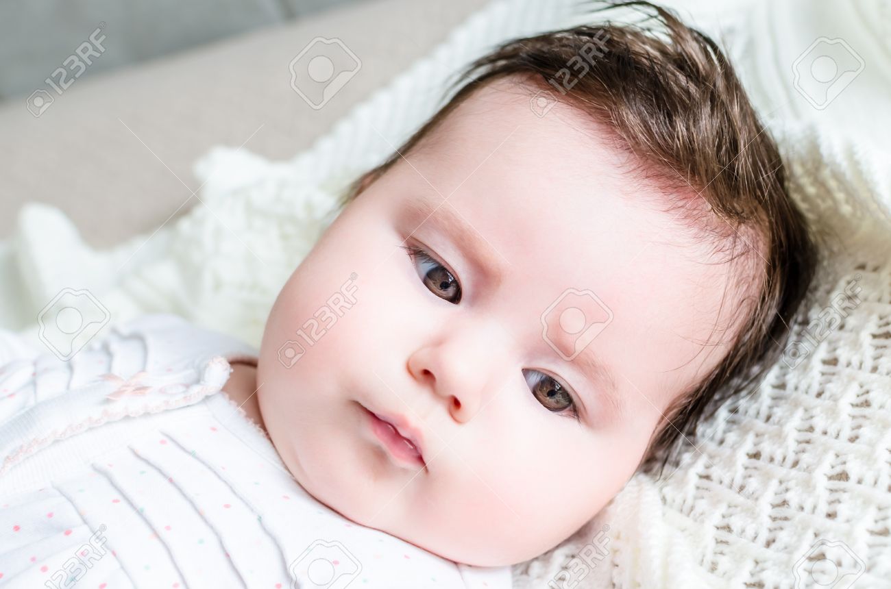 Retrato De La Nina Lindo Bebe Recien Nacido Dulce Morena Con El Pelo Negro Que Mira Con Grandes Hermosa Avellana Ojos Marrones Y Acostado Sobre Una Manta De Lana Blanca En Una