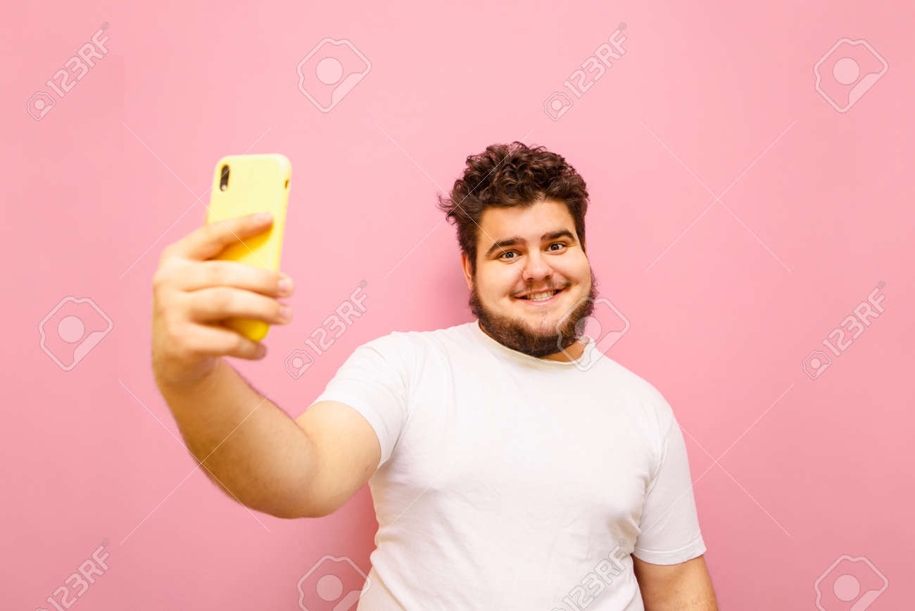 Funny Fat Guy With Curly Hair Takes A Selfie On A Pink Background, Looks Into The Funny Fat Guy With Curly Hair Takes A Selfie On A Pink Background, Looks Into The