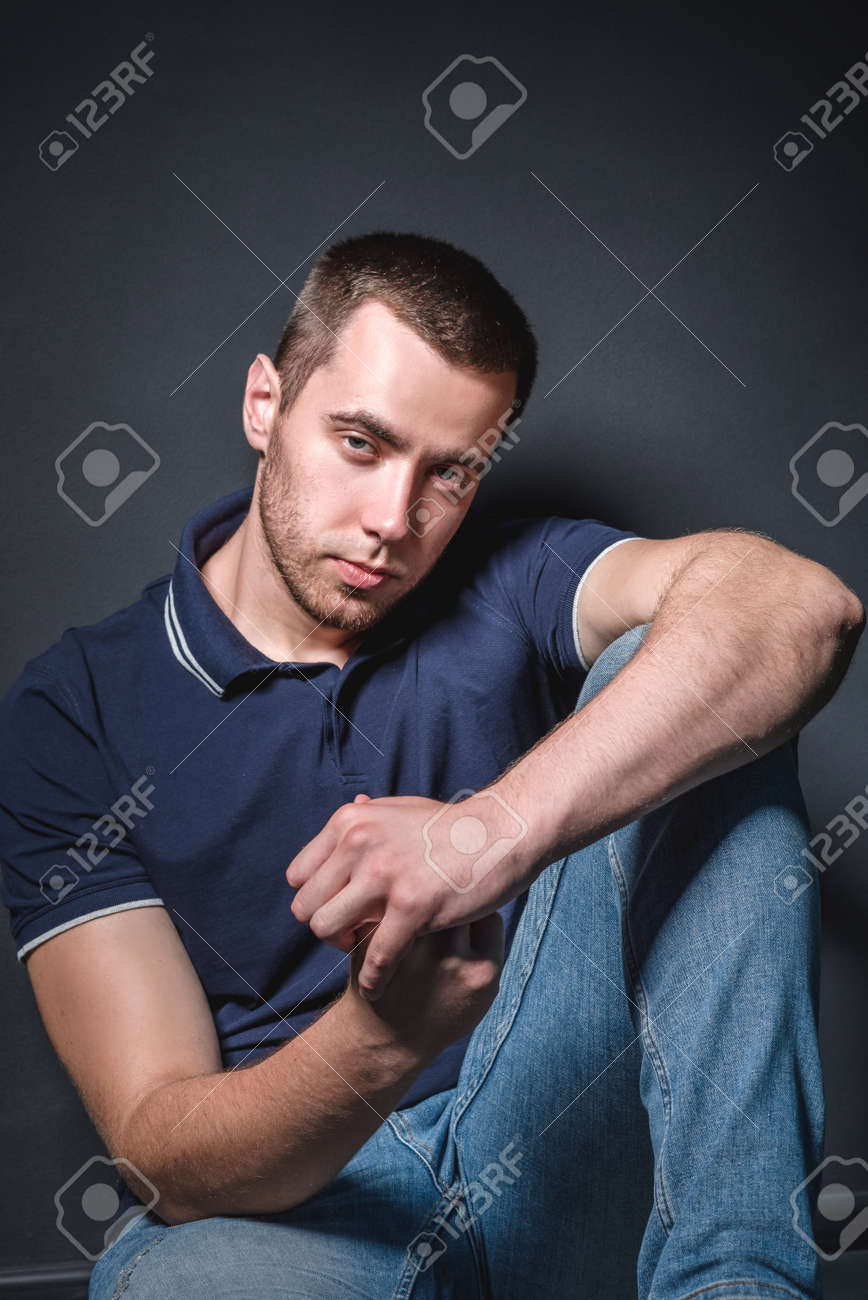 Young attractive man in blue jeans and a polo shirt sits on the floor near the wall in the studio. Youth and attractiveness concept - 155813226 Young attractive man in blue jeans and a polo shirt sits on the floor near the wall in the studio. Youth and attractiveness concept - 155813226