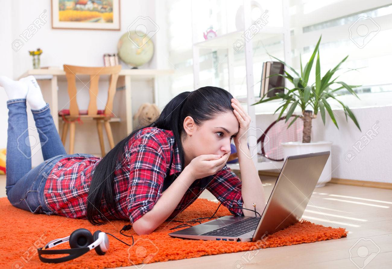 Young girl lying on the carpet in room and looking at laptop with shocked expression - 57148078 Young girl lying on the carpet in room and looking at laptop with shocked expression - 57148078