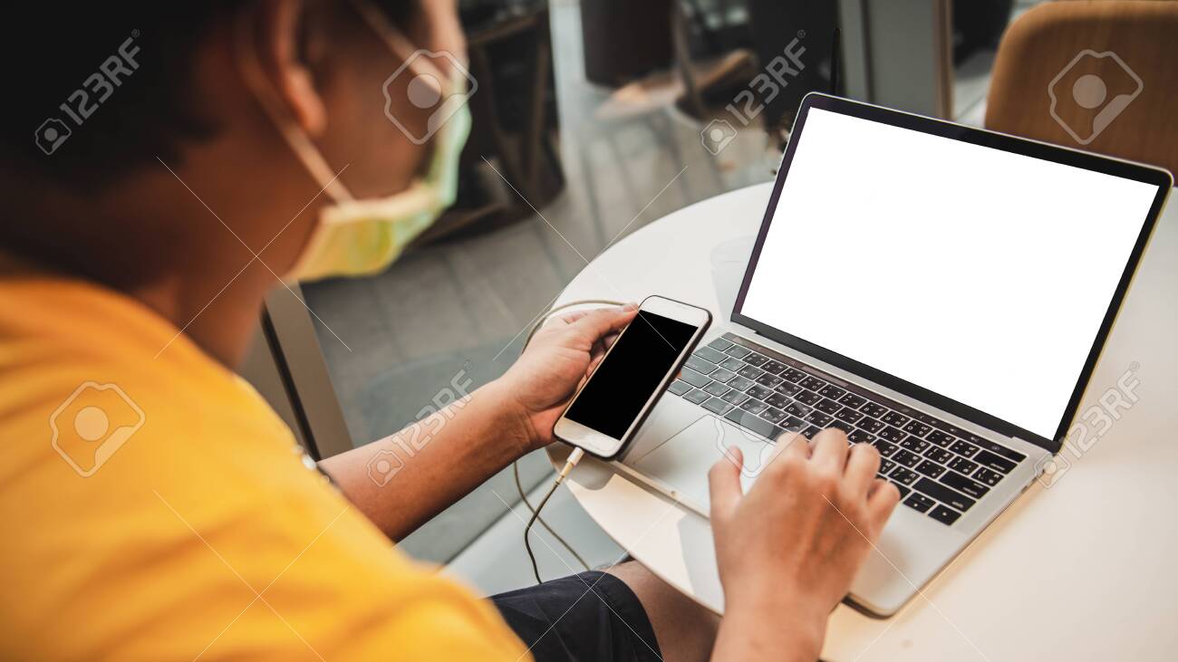 A man in yellow shirt and medical mask using laptop and smartphone while sitting in coffeeshop - 148652046 A man in yellow shirt and medical mask using laptop and smartphone while sitting in coffeeshop - 148652046