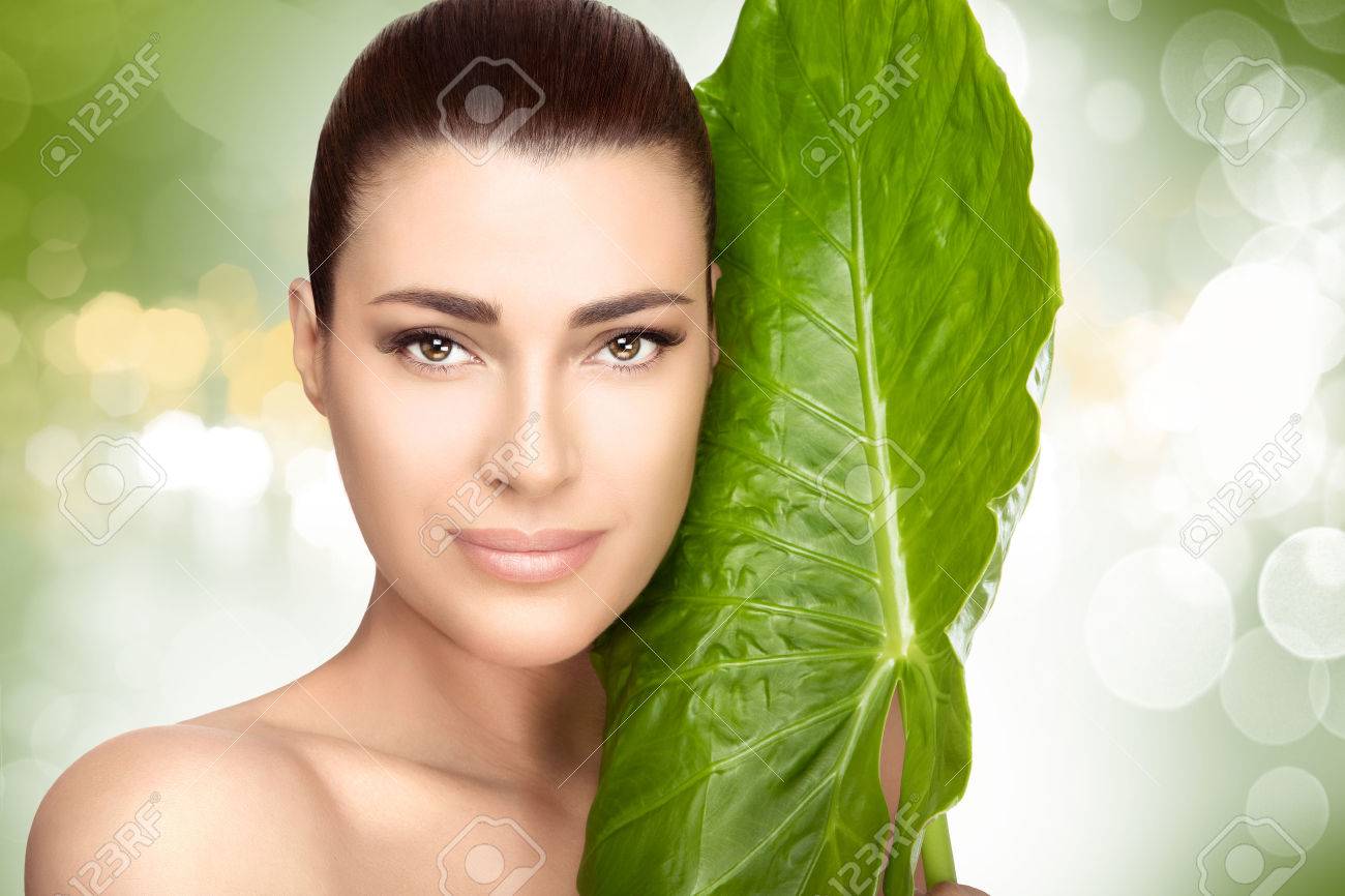 Beauty portrait of an attractive natural young girl with a large fresh green leaf held to her cheek against a soft green bokeh background in a spa and wellness concept - 54110300 Beauty portrait of an attractive natural young girl with a large fresh green leaf held to her cheek against a soft green bokeh background in a spa and wellness concept - 54110300
