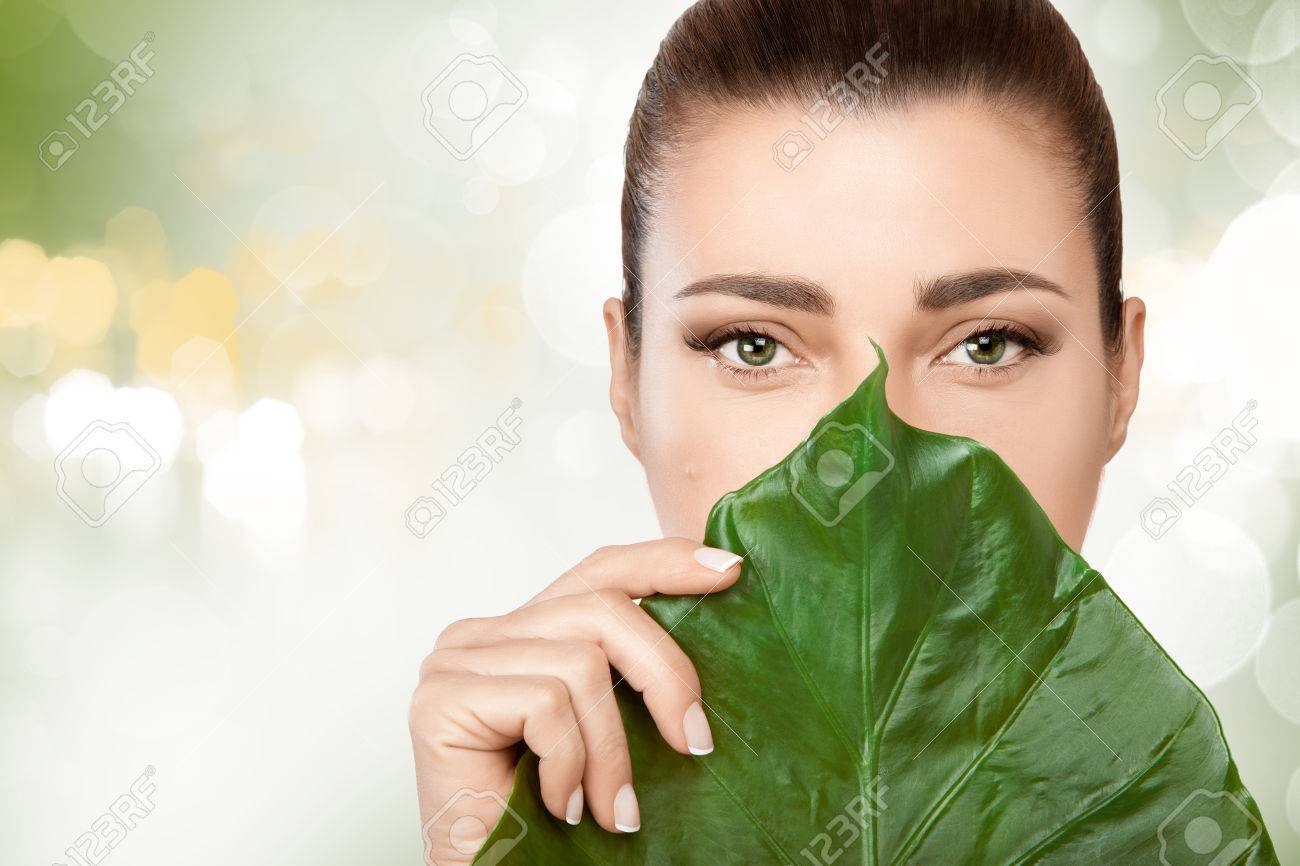 Gorgeous young brunette woman with a fresh leaf from a tropical plant held to her face covering mouth and nose in a spa and beauty concept. Portrait over soft green bokeh background with copy space - 54110296 Gorgeous young brunette woman with a fresh leaf from a tropical plant held to her face covering mouth and nose in a spa and beauty concept. Portrait over soft green bokeh background with copy space - 54110296