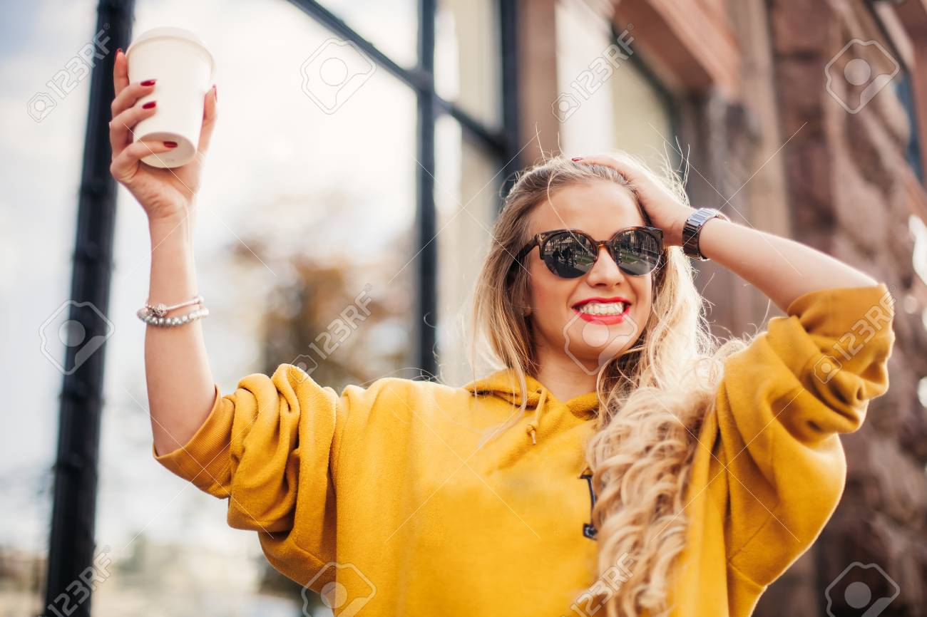 Stylish happy young woman wearing boyfrend jeans, white sneakers bright yellow sweetshot.She holds coffee to go. portrait of smiling girl in sunglasses posing in the street - 89978268 Stylish happy young woman wearing boyfrend jeans, white sneakers bright yellow sweetshot.She holds coffee to go. portrait of smiling girl in sunglasses posing in the street - 89978268