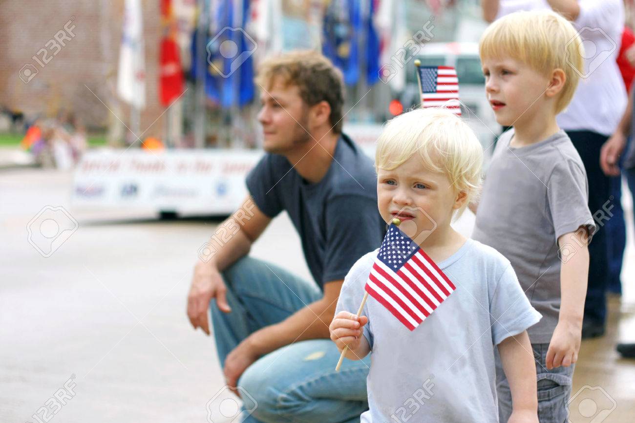 Two young boy children are waving American Flags as they stand with their Father at watch a Patriotic Parade Event in the United States. - 65013928 Two young boy children are waving American Flags as they stand with their Father at watch a Patriotic Parade Event in the United States. - 65013928