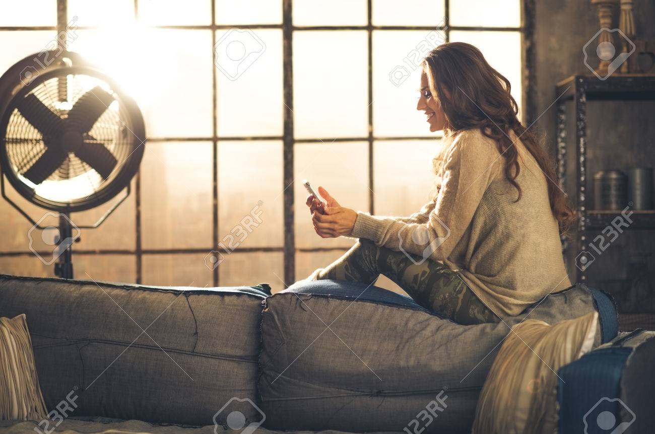 Seen from the side,a brunette woman is smiling, looking down at her phone sitting on the back of a sofa. Industrial chic ambiance and cozy atmosphere, sunlight is streaming through the loft window. - 40213899 Seen from the side,a brunette woman is smiling, looking down at her phone sitting on the back of a sofa. Industrial chic ambiance and cozy atmosphere, sunlight is streaming through the loft window. - 40213899