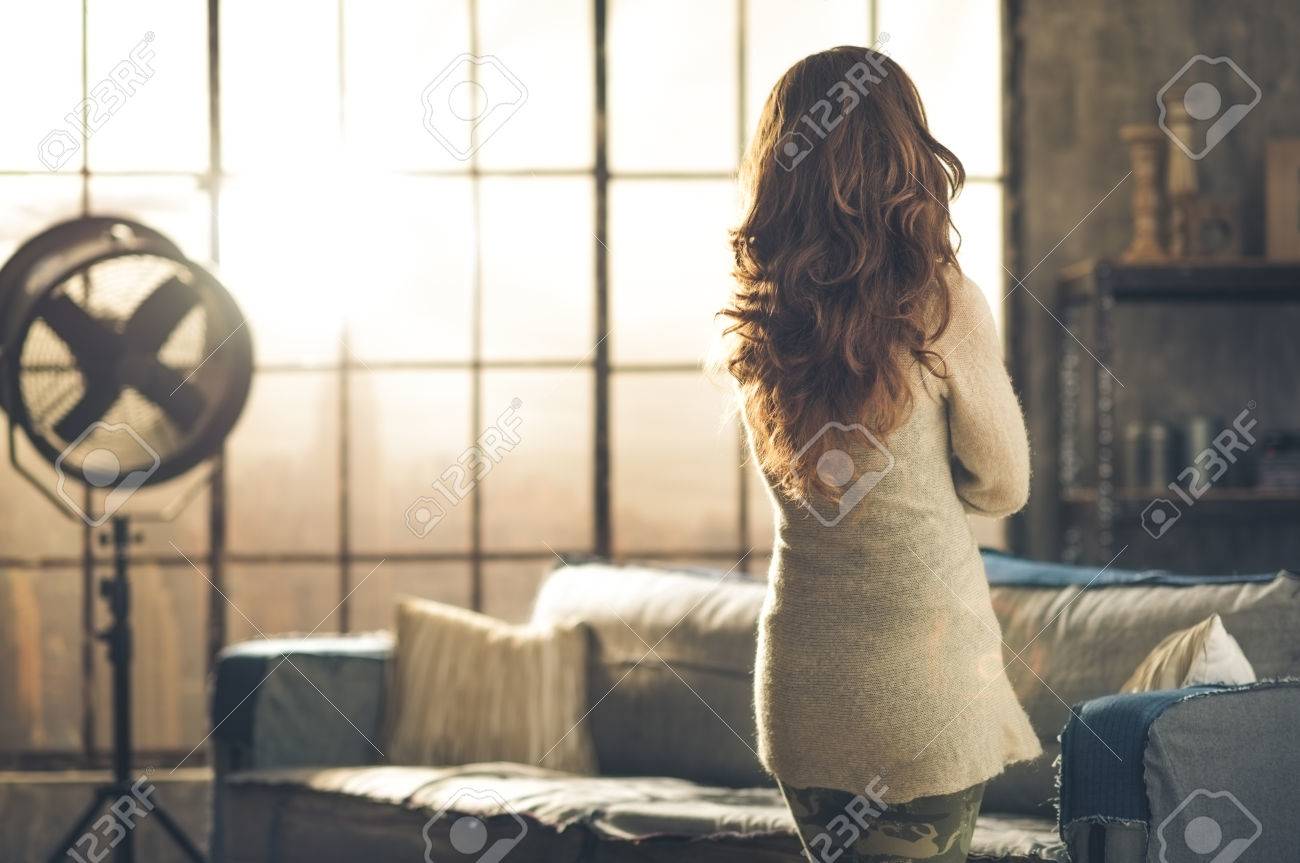 Seen from behind, a brunette woman in comfortable clothing is standing in a loft living room, looking out the loft window. Urban chic loft decoration details. - 40186473 Seen from behind, a brunette woman in comfortable clothing is standing in a loft living room, looking out the loft window. Urban chic loft decoration details. - 40186473