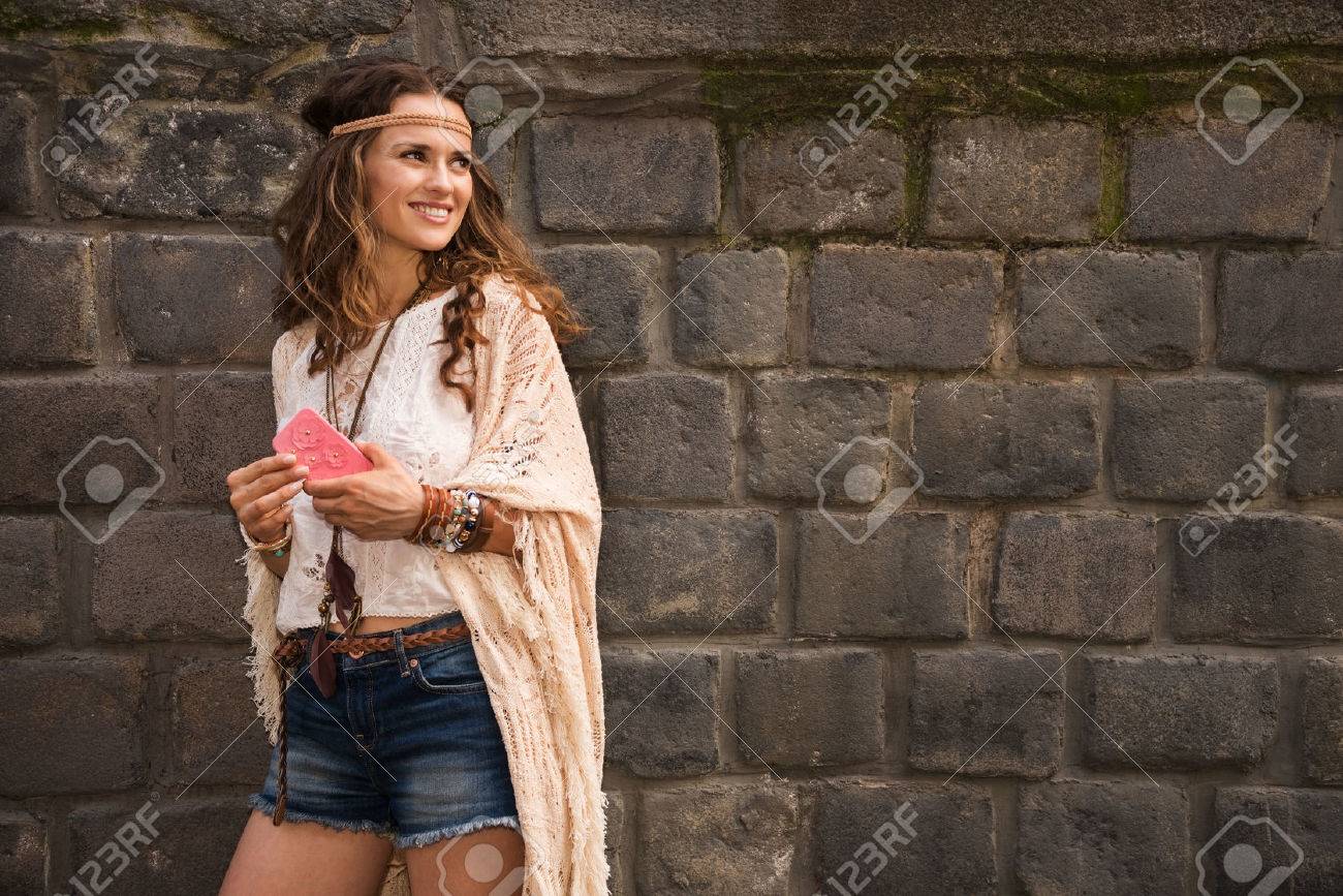 Longhaired hippy-looking young lady in jeans shorts, knitted shawl and white blouse standing near stone wall in old town and holding cell phone - 43887953 Longhaired hippy-looking young lady in jeans shorts, knitted shawl and white blouse standing near stone wall in old town and holding cell phone - 43887953