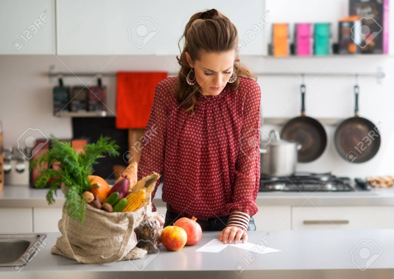 An elegant woman looks down at the shopping lists on her kitchen counter, reading them carefully. Next to her on the kitchen counter, a burlap sac holds a wide variety of fall fruits and vegetables. - 45387390 An elegant woman looks down at the shopping lists on her kitchen counter, reading them carefully. Next to her on the kitchen counter, a burlap sac holds a wide variety of fall fruits and vegetables. - 45387390