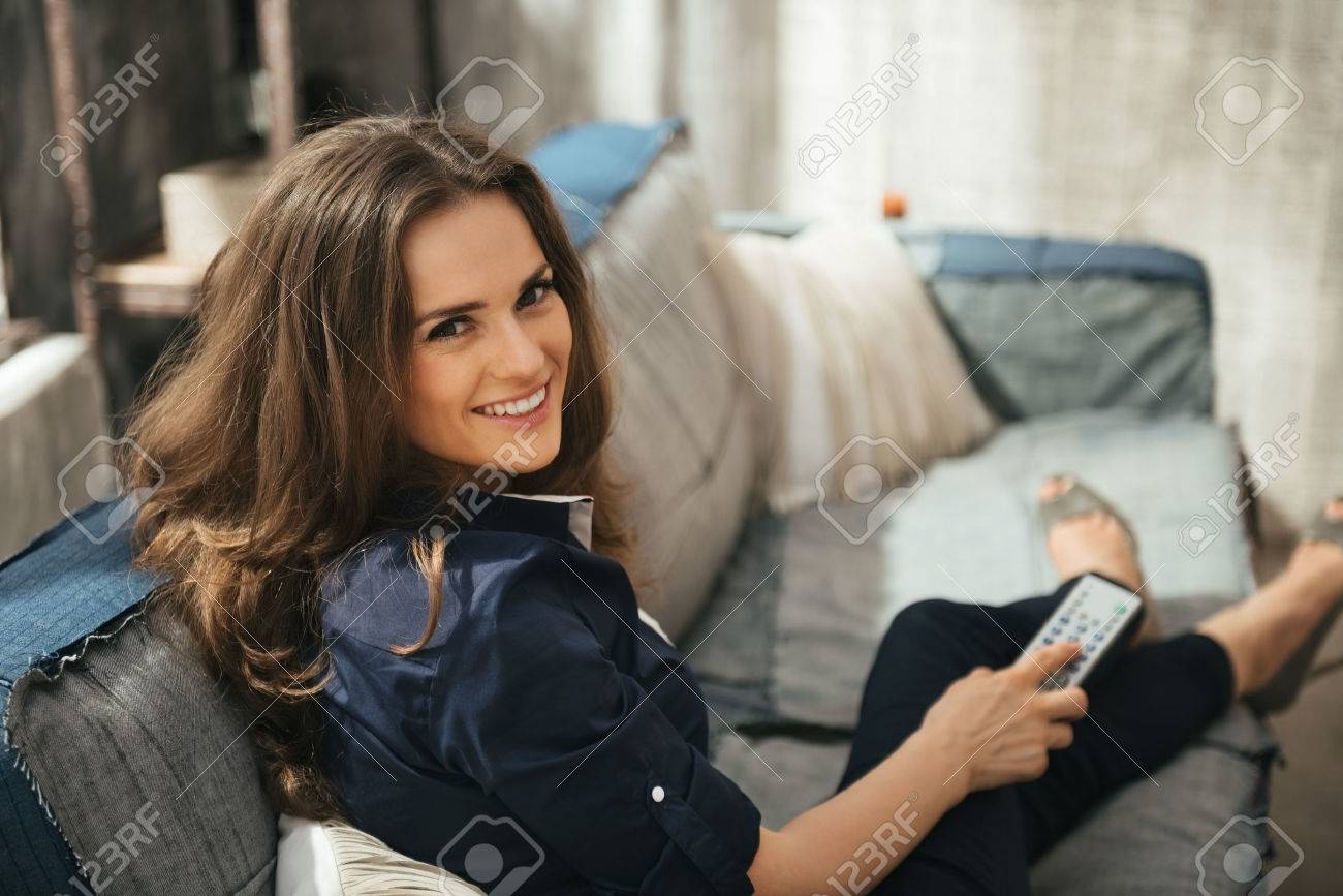 Smiling longhaired woman with TV remote control in hand relaxing on sofa and watching tv in loft apartment. Urban chic loft decoration details and window. Modern lifestyle concept. - 46668580 Smiling longhaired woman with TV remote control in hand relaxing on sofa and watching tv in loft apartment. Urban chic loft decoration details and window. Modern lifestyle concept. - 46668580
