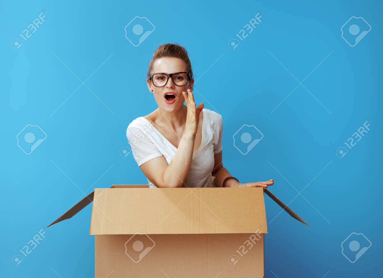 young woman in white t-shirt in a cardboard box telling exciting news isolated on blue background - 116502056 young woman in white t-shirt in a cardboard box telling exciting news isolated on blue background - 116502056