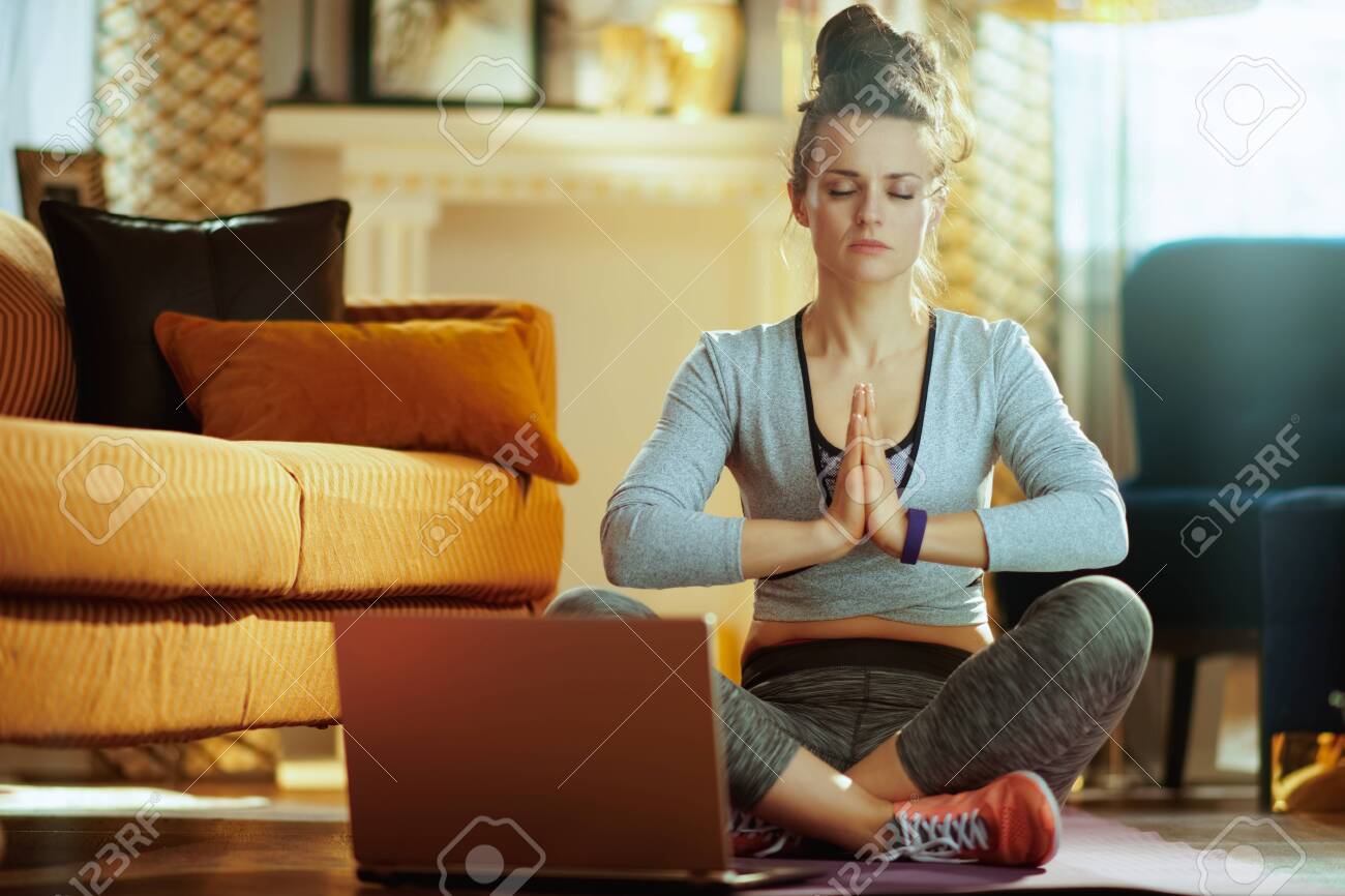 relaxed fit sports woman in fitness clothes in the modern house meditating using online yoga training program in laptop. - 121842294 relaxed fit sports woman in fitness clothes in the modern house meditating using online yoga training program in laptop. - 121842294