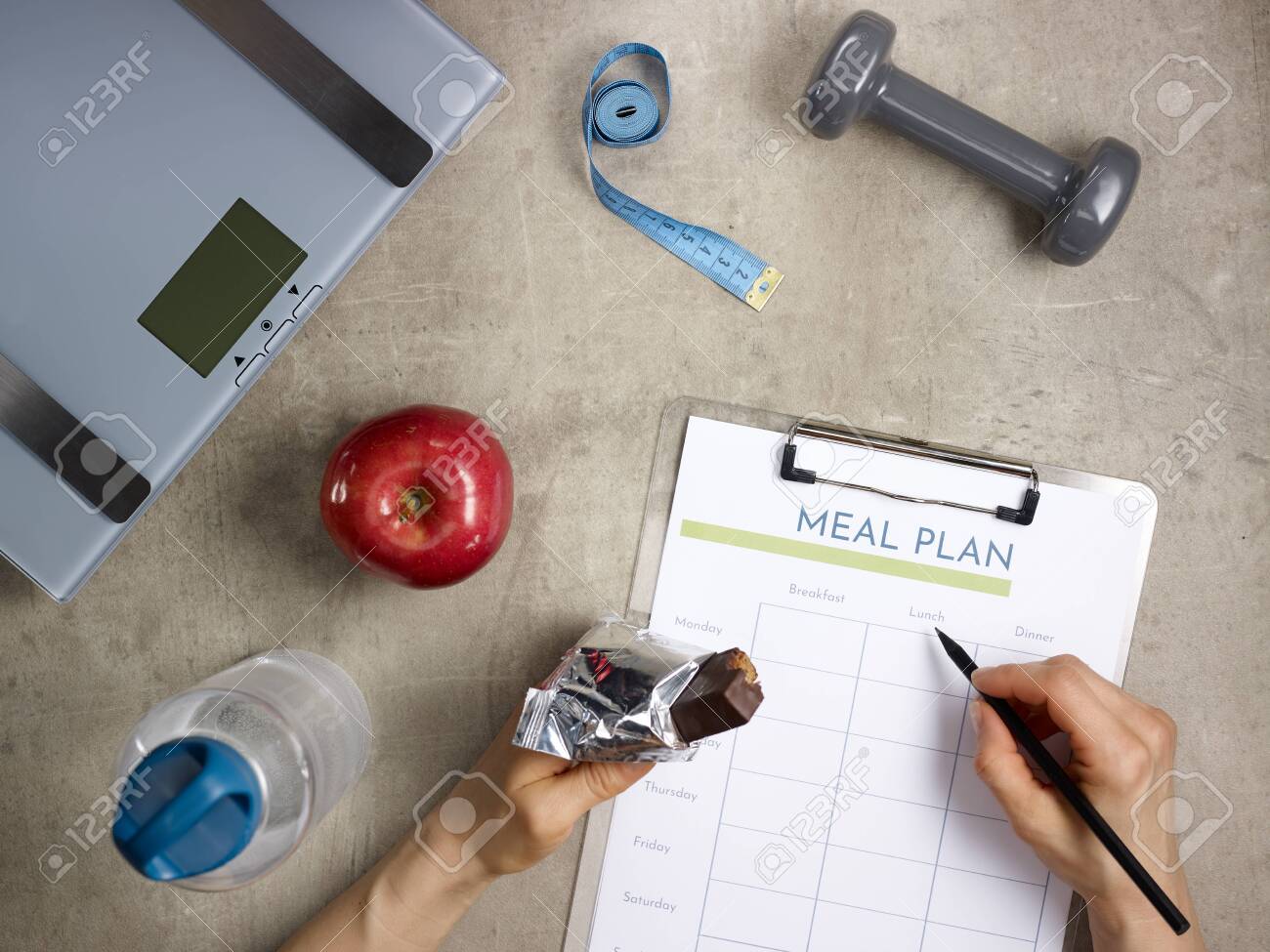 Closeup on weight scales, grey dumbbell, red apple, bottle of water, tape measure laying on the floor and female hands with bitten raw protein bar filling meal plan. - 122620568 Closeup on weight scales, grey dumbbell, red apple, bottle of water, tape measure laying on the floor and female hands with bitten raw protein bar filling meal plan. - 122620568