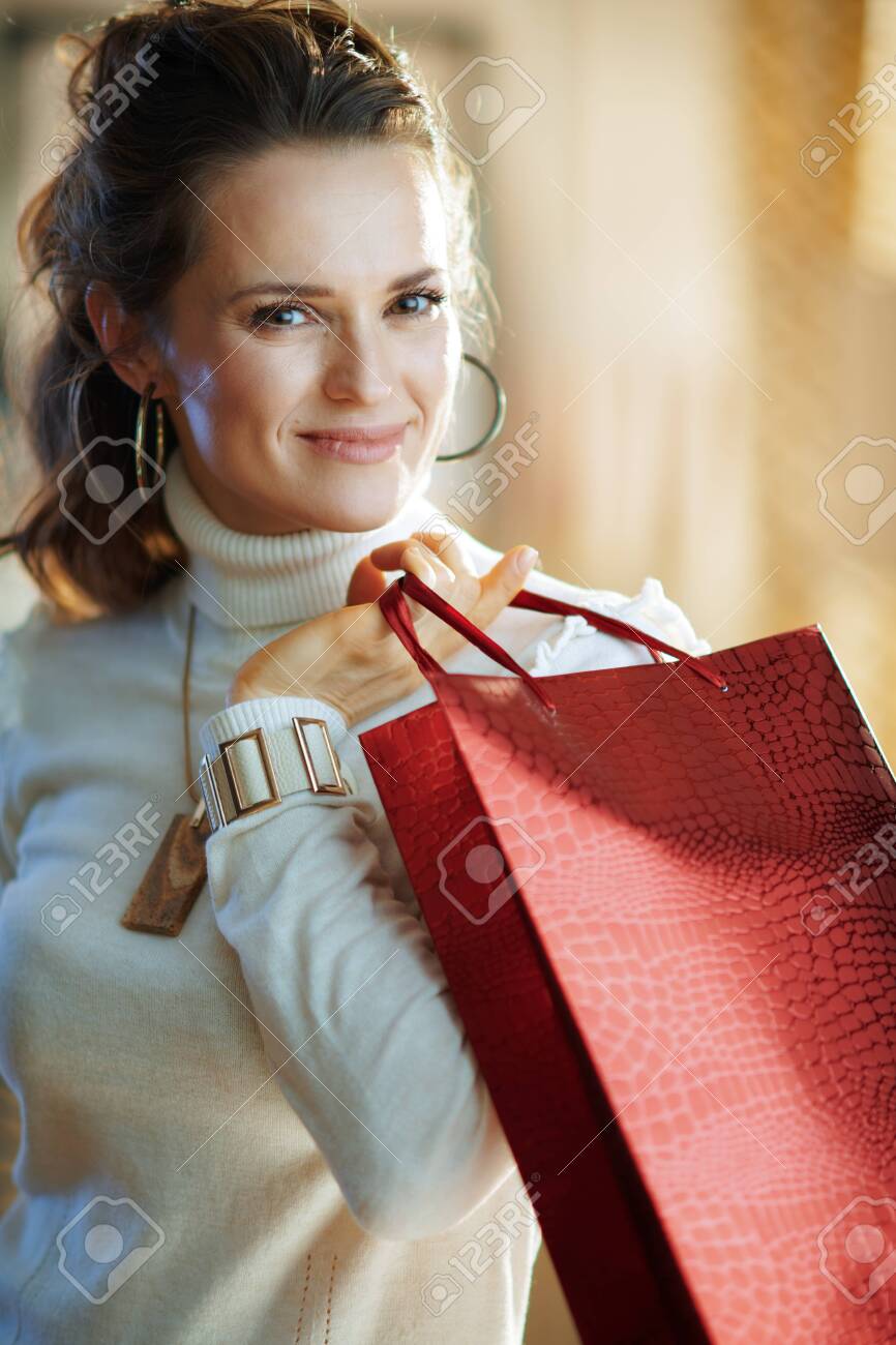 Portrait of elegant female in white sweater and skirt with red shopping bag in the modern house in sunny winter day. - 140598984 Portrait of elegant female in white sweater and skirt with red shopping bag in the modern house in sunny winter day. - 140598984