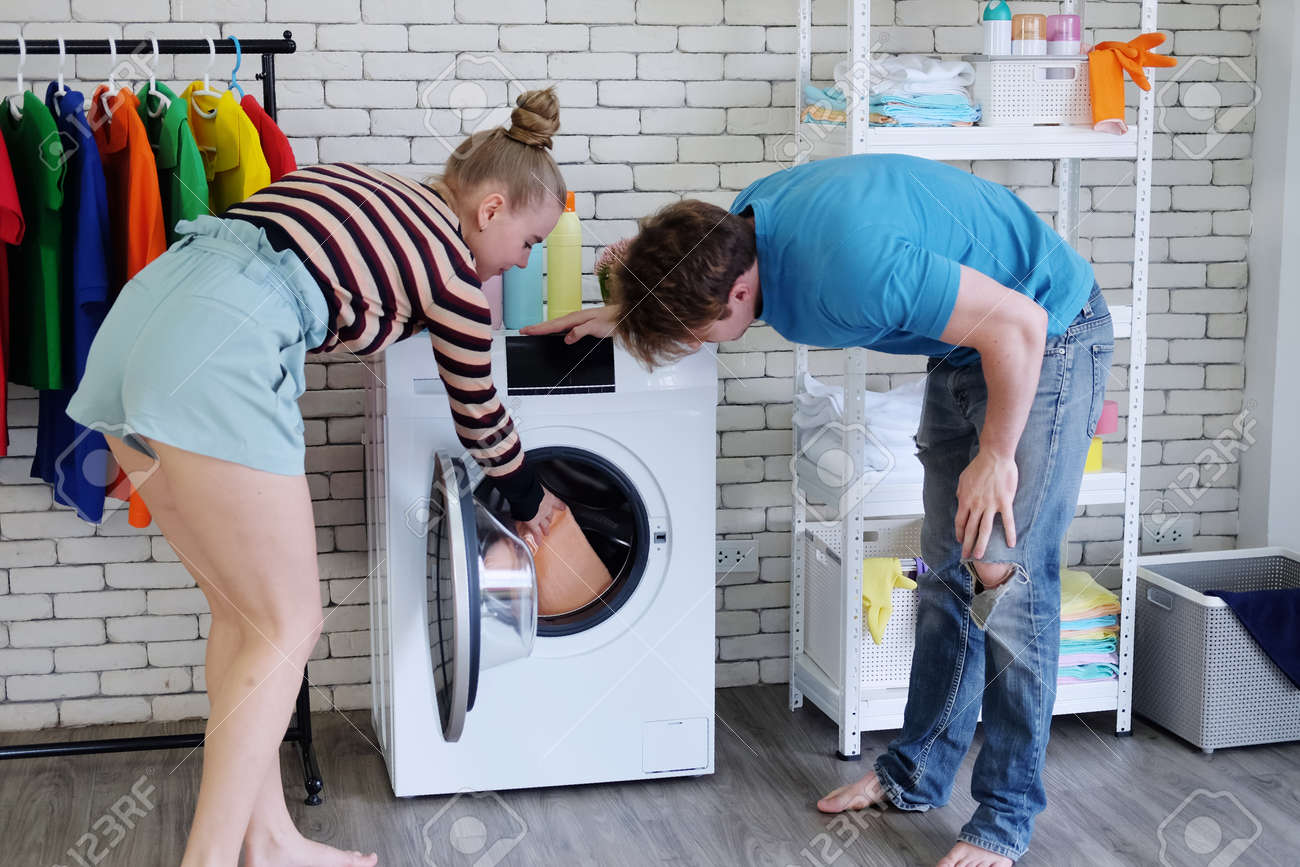 Smiling Young Caucasian Couple Love Husband And Wife Is Pick Up The Clothes  In The Washing Machine Together In Laundry Room. Lifestyle Of Family And  Married On Holiday Concept Stock Photo, Picture