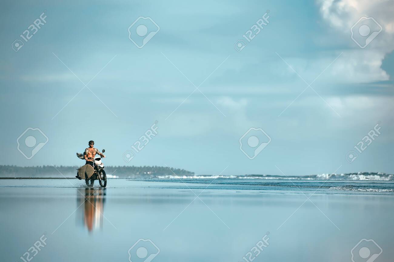 Authentic male surfer rides with surfboard on motorcycle at ocean beach. Real Bali surfing photo - 144779003 Authentic male surfer rides with surfboard on motorcycle at ocean beach. Real Bali surfing photo - 144779003