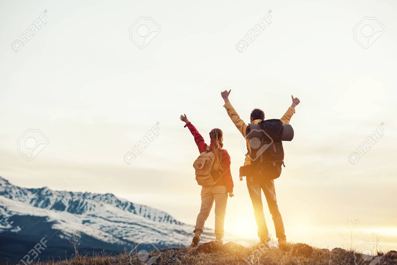 Two happy hikers with backpacks are standing with raised arms and looking at mountains and sunset - 145683326 Two happy hikers with backpacks are standing with raised arms and looking at mountains and sunset - 145683326