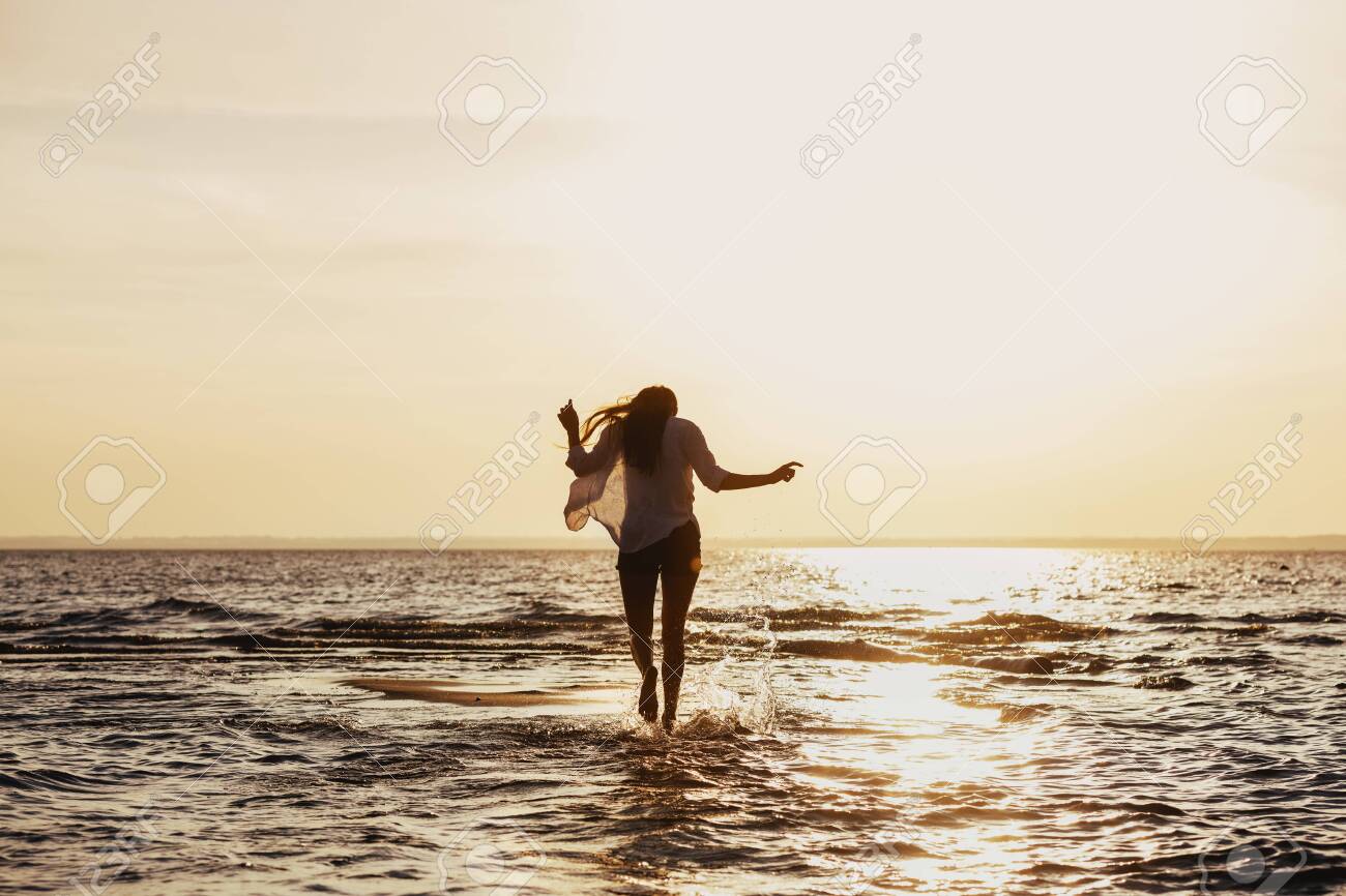Young slim girl walks by water at sunset beach - 146342079 Young slim girl walks by water at sunset beach - 146342079