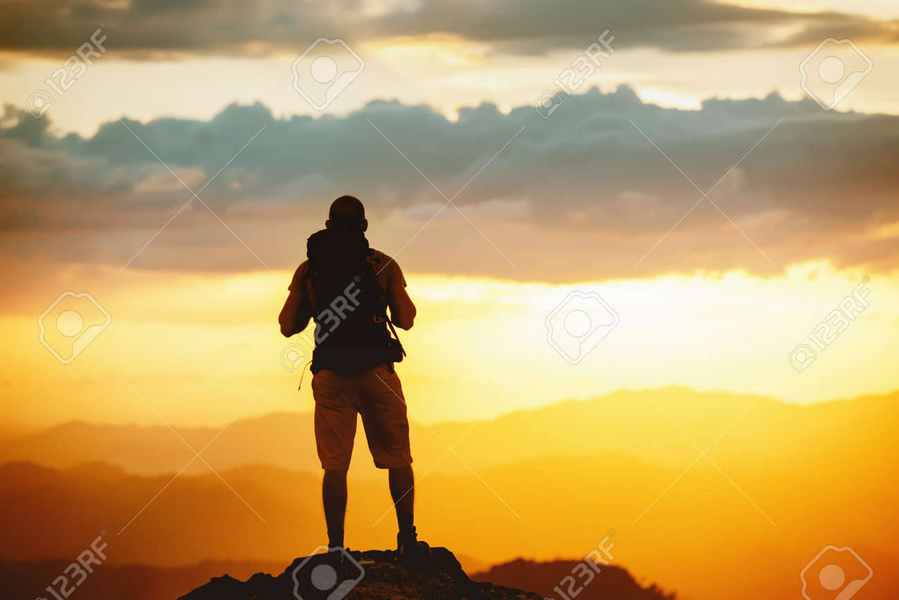 Unrecognized hiker stands with backpack on mountain top and enjoys sunset sky - 154759377 Unrecognized hiker stands with backpack on mountain top and enjoys sunset sky - 154759377