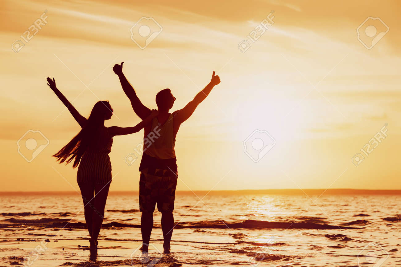 Happy couple stands with raised arms at sunset beach - 159457852 Happy couple stands with raised arms at sunset beach - 159457852