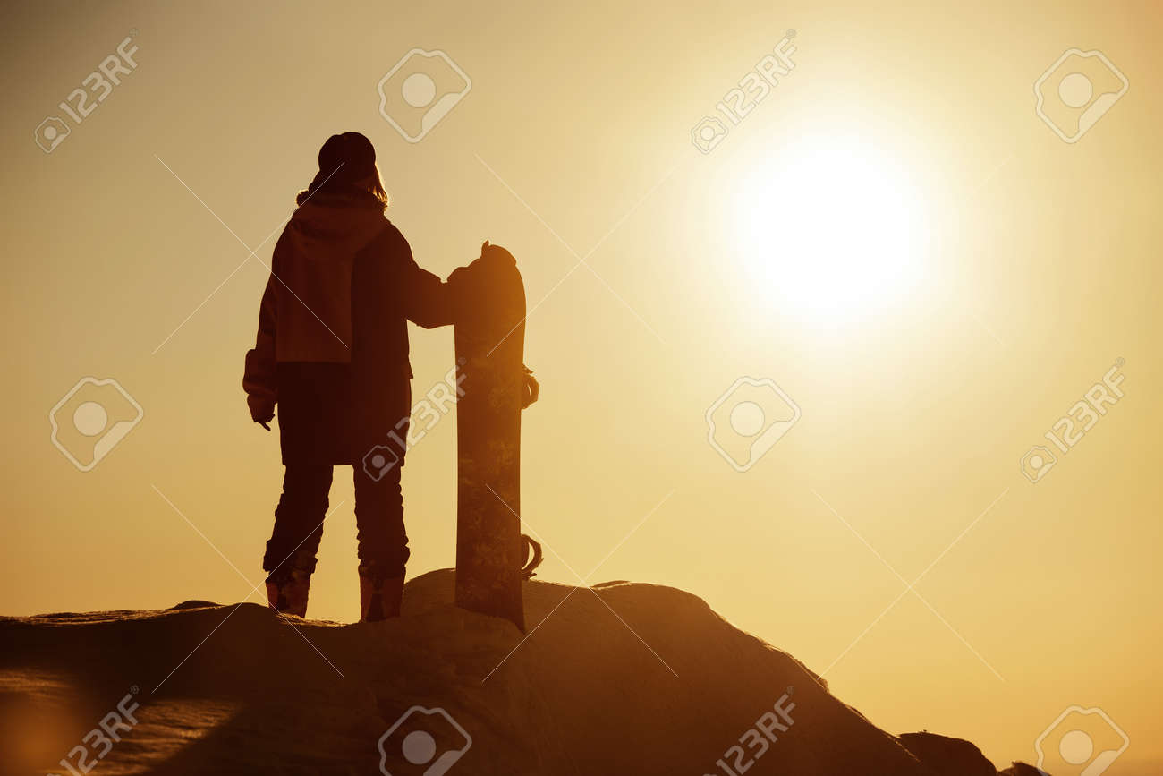 Girl snowboarder stands with snowboard against sunset sky. Ski resort concept - 159685013 Girl snowboarder stands with snowboard against sunset sky. Ski resort concept - 159685013