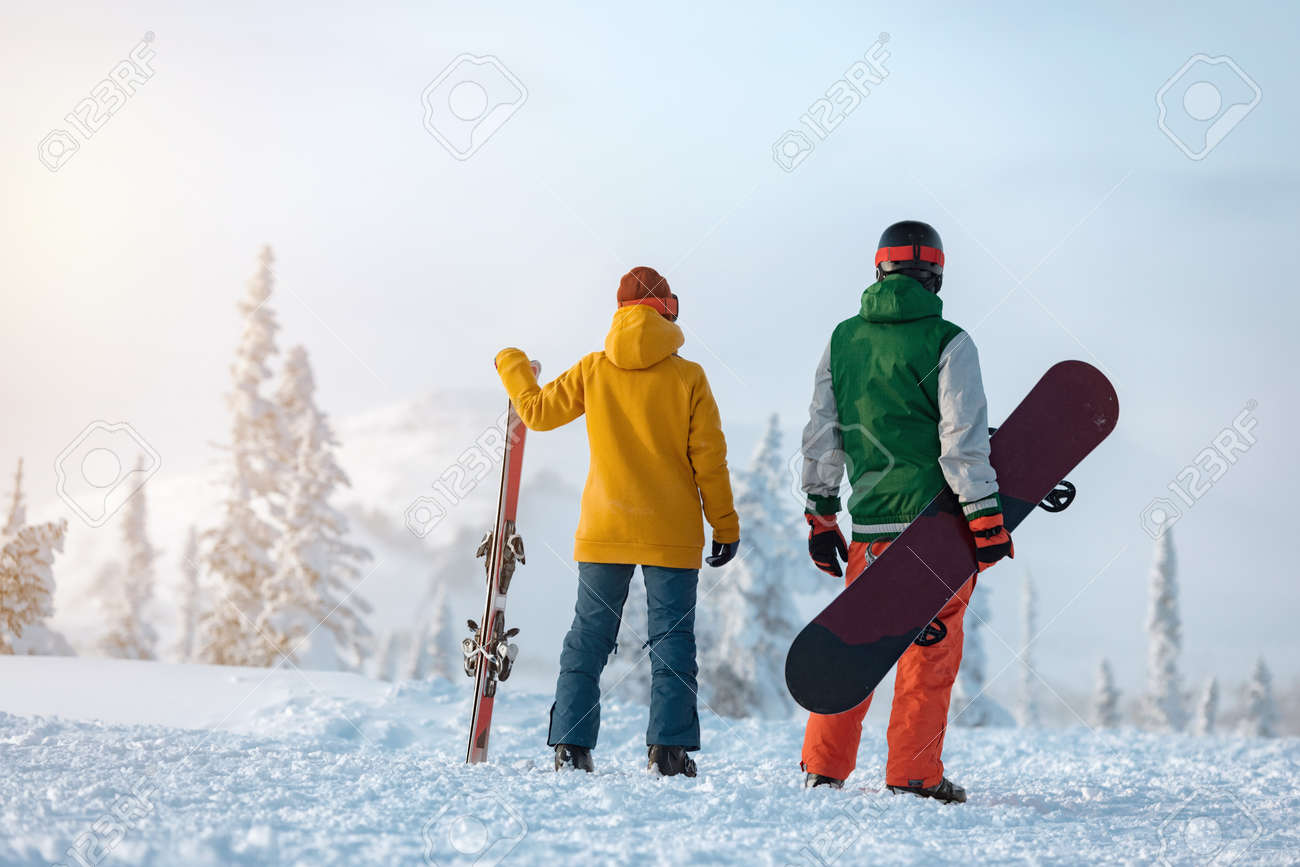 Female skier and male snowboarder are standing on background of snowcapped mountain at sunset time. Ski resort concept - 165555399 Female skier and male snowboarder are standing on background of snowcapped mountain at sunset time. Ski resort concept - 165555399