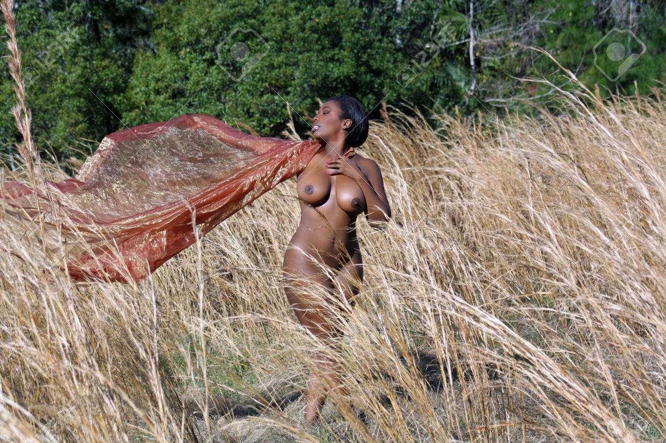 A lovely, sexy, nude, black woman standing in tall grass, holding..