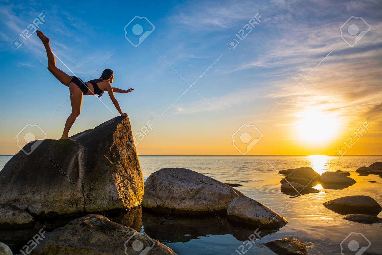 Full length slim female balancing on rock in Virabhadrasana pose against sunset sky during yoga lesson near sea - 163148240 Full length slim female balancing on rock in Virabhadrasana pose against sunset sky during yoga lesson near sea - 163148240