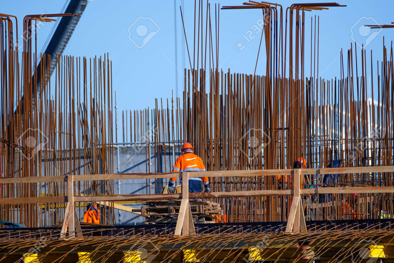 Anonymous men in uniform working on bridge near metal poles outside unfinished building on sunny day on construction site - 166802195 Anonymous men in uniform working on bridge near metal poles outside unfinished building on sunny day on construction site - 166802195