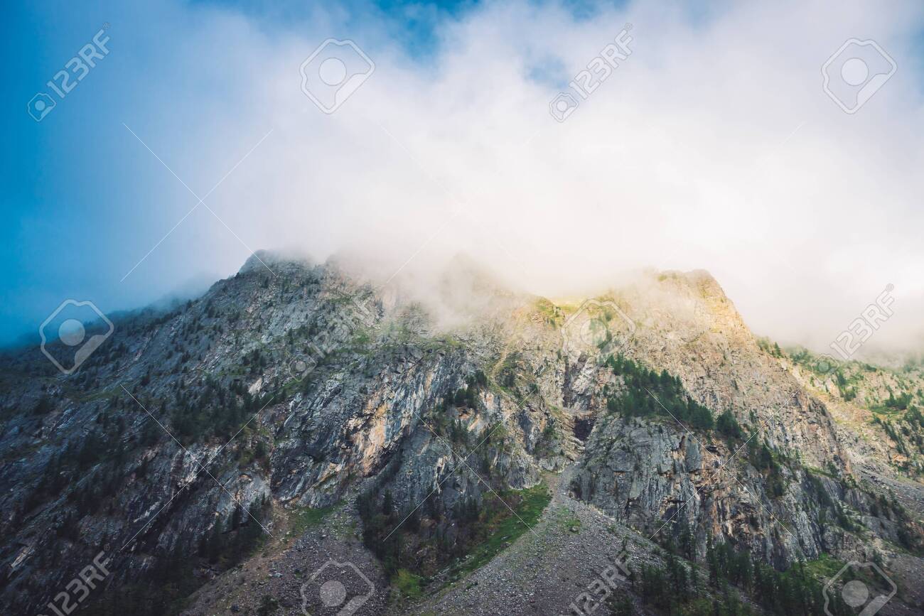 Giant cloud above rocky ridge with trees in sunlight. Amazing mountain range under blue cloudy dawn sky. Wonderful foggy rocks. Misty landscape of highland nature. Low clouds in morning sunny light. - 122474920 Giant cloud above rocky ridge with trees in sunlight. Amazing mountain range under blue cloudy dawn sky. Wonderful foggy rocks. Misty landscape of highland nature. Low clouds in morning sunny light. - 122474920