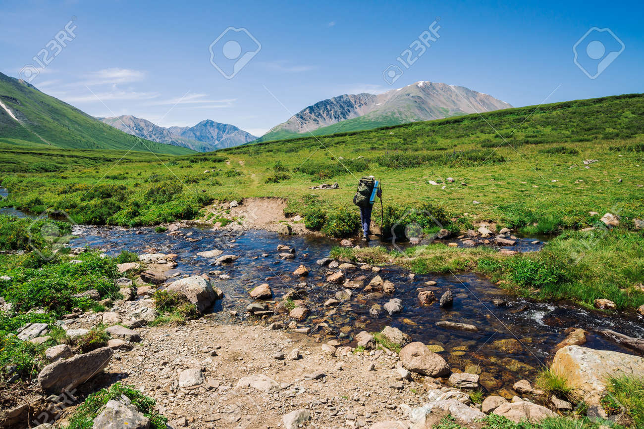 Man with large backpack crosses mountain creek over stones. Tourist goes on path in green valley to wonderful snowy mountains. Hiking in highlands. Amazing atmospheric landscape of majestic nature. - 122478620 Man with large backpack crosses mountain creek over stones. Tourist goes on path in green valley to wonderful snowy mountains. Hiking in highlands. Amazing atmospheric landscape of majestic nature. - 122478620