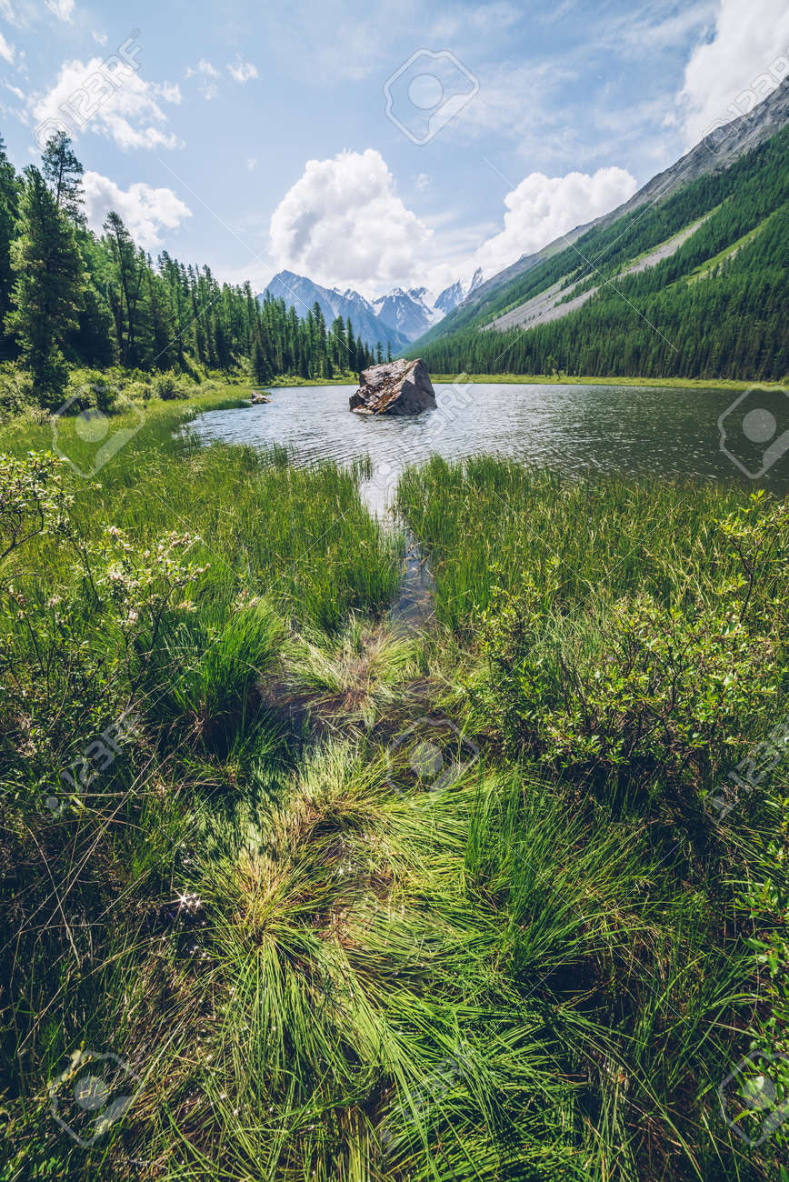 Meditative view to beautiful lake with stone in valley on snowy mountains background. Scenic relaxing green landscape with big mossy stone in mountain lake. Alpine lake with ripples on water surface. - 170160615 Meditative view to beautiful lake with stone in valley on snowy mountains background. Scenic relaxing green landscape with big mossy stone in mountain lake. Alpine lake with ripples on water surface. - 170160615