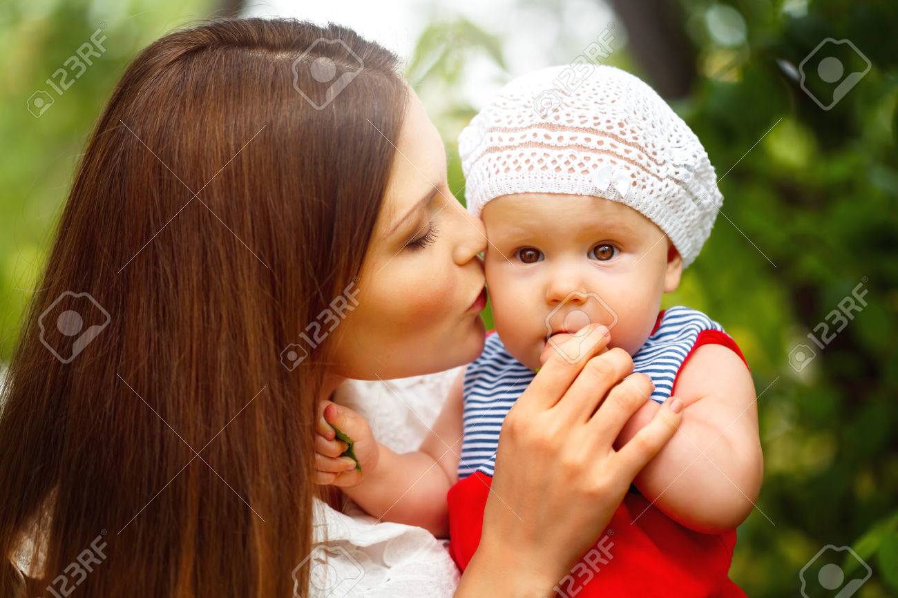 カメラに見て ママの手の上に座って驚いた幼児の赤ちゃん女の子 母親は子供にキスします 緑の夏公園でリラックスし 緑の背景がぼやけています クローズ アップの肖像画 セレクティブ フォーカス の写真素材 画像素材 Image