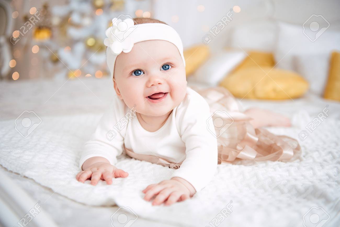 Baby girl wearing cute dress and headband, lies on a white cover in festively decorated room. With surprise watches in the camera, on a background a set of bright fires, soft focus. Warm beige and gold colors - 92058645 Baby girl wearing cute dress and headband, lies on a white cover in festively decorated room. With surprise watches in the camera, on a background a set of bright fires, soft focus. Warm beige and gold colors - 92058645
