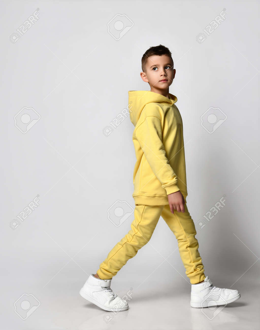 Vista Lateral De Un Niño Que Camina Un Chándal Amarillo Con Capucha Y Pantalones, Zapatillas Blancas, Aislado En Blanco. Retrato De Un Niño Con Ropa Fresca. Moda Infantil, Casual, Ropa